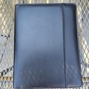 May include: A black leather portfolio with a stitched border and a vertical seam. The portfolio is rectangular and appears to be closed. The surface has a slight sheen, suggesting a polished finish. The background is a metal grate.