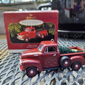 May include: A red 1953 GMC All-American truck ornament with a Christmas tree in the bed. The ornament is a Hallmark Keepsake. The truck has black tires and red rims. The box is red with an image of the truck.