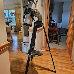 May include: A silver and black telescope mounted on a black tripod stand. The telescope features a cylindrical optical tube with an eyepiece at the top, a side-mounted finder scope, and a circular adjustment knob on the side of the mounting arm.