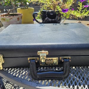 May include: Black briefcase with gold-colored hardware, including a combination lock and clasps. The handle is black leather. The briefcase is sitting on a metal table.