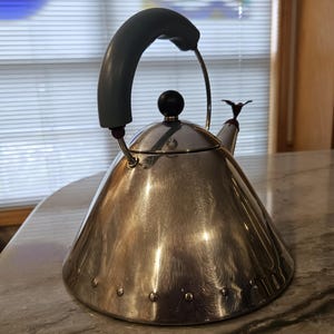May include: A conical stainless steel tea kettle with a polished metallic finish. It features a gray arched handle, a black spherical lid knob, and a small bird-shaped whistle attachment on the spout. A row of small rivets circles the base.