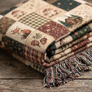 May include: A woven throw blanket featuring a patchwork design of squares with various patterns including mushrooms, a cabin in the woods, a fox, acorns, and green plaid. The blanket has a fringed edge with multi-coloured threads along the bottom.