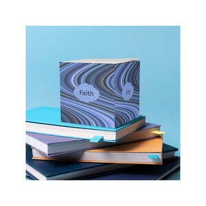 May include: A cube-shaped decorative item with a swirling blue and white pattern, labeled "Faith" and "Joy", sits atop a stack of colorful books. The books have blue, purple, and orange covers with blue and yellow page markers.