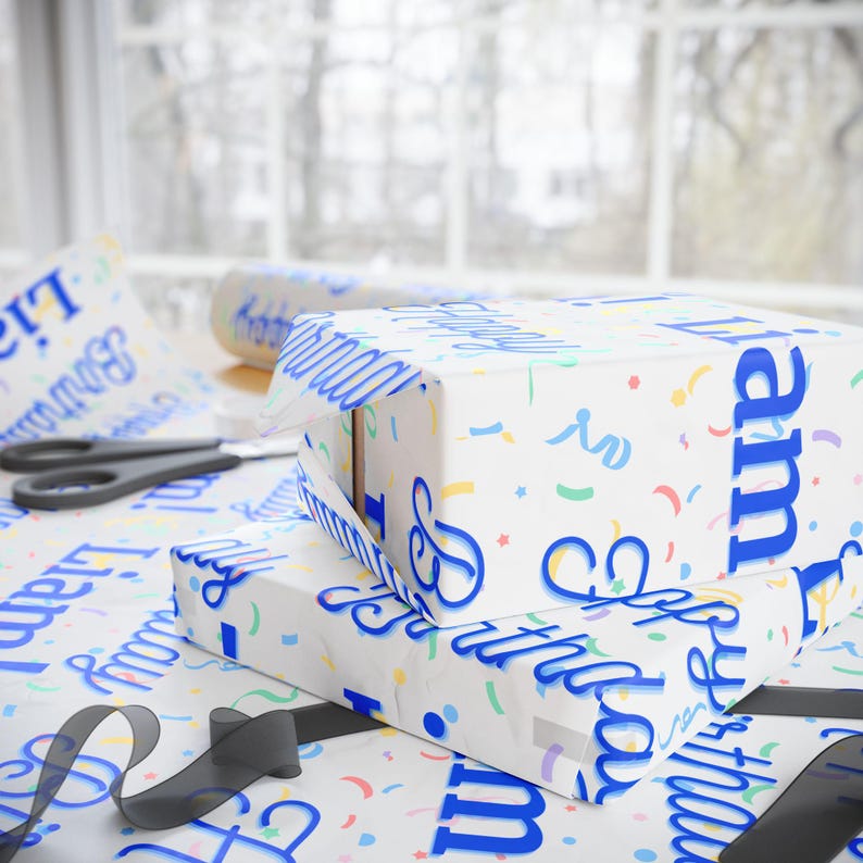 Two wrapped gifts with "Happy Birthday Liam" paper and a roll of matching paper are on a white table with scissors and black ribbon.