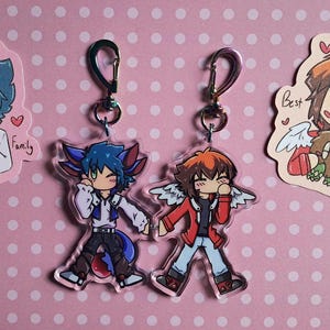 May include: Acrylic anime character keychains and stickers. The keychains feature two characters with wings, and the stickers show anime characters with text that says "My Precious Family" and "Best Bud".