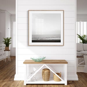 May include: A framed black and white photograph of the ocean hangs above a wooden console table. The table features a light wood top, white base, and decorative items, including a glass bowl and woven baskets. The room has white walls and hardwood floors.