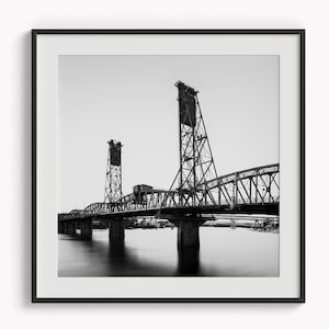 May include: Black and white photograph of a steel bridge over a body of water. The bridge has two towers and is made of metal beams. The water is calm and reflects the sky.