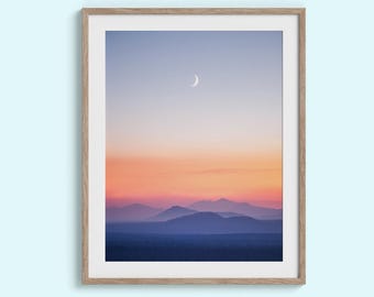 May include: A framed landscape print featuring a serene mountain range under a gradient sky. The sky transitions from a soft blue to vibrant orange and pink hues, with a crescent moon visible. The mountains are a deep blue.