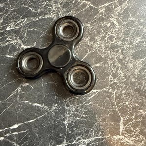 May include: A black fidget spinner with three rounded arms and circular bearings. The center of the spinner has a circular cap. The spinner is set against a dark, marbled background.