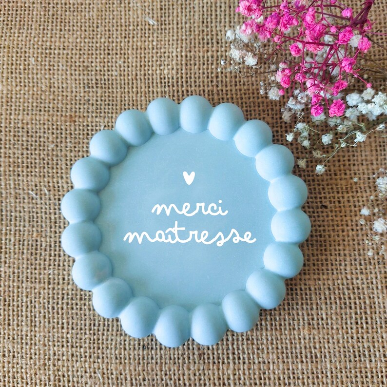 May include: A light blue, round decorative plate with a scalloped edge. The plate features the text "merci ma&icirc;tresse" in white script, along with a small white heart. Pink and white flowers are in the background.