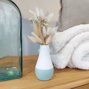 Op de afbeelding: Een kleine, witte en lichtblauwe keramische vaas met een boeket gedroogde witte pampasgras en witte baby's breath.