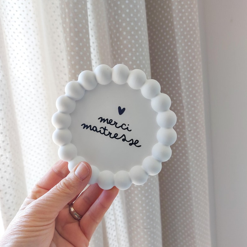 May include: A white, round coaster with a beaded edge. The coaster has the words "merci ma&icirc;tresse" in black script with a small heart above the text. The coaster is held in a hand.