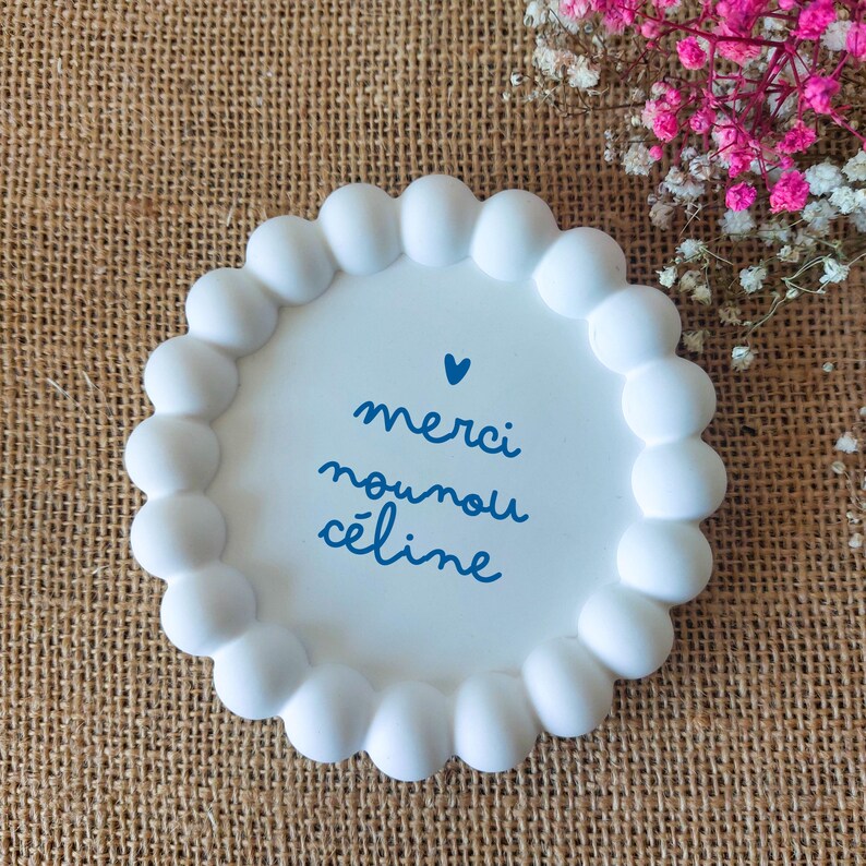 May include: A white, round decorative tray with a scalloped edge. The tray features blue script that reads "merci nounou c&eacute;line" with a small blue heart. The tray is on a textured, brown surface.