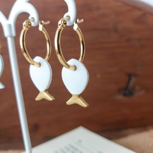 May include: Gold hoop earrings featuring white fish-shaped pendants with gold-coloured tails. The earrings are displayed on a white stand. The fish pendants have a minimalist design, set against a blurred background.