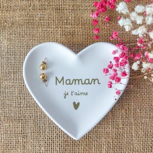 Peut inclure: Un vide-poche blanc en forme de cœur avec l'inscription "Maman je t'aime" en vert. Deux boucles d'oreilles dorées sont posées sur le plat. Des fleurs roses et blanches sont en arrière-plan. Le plat est sur une surface en toile de jute.