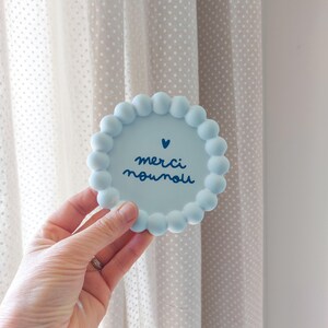 May include: A light blue, round coaster with a beaded edge. The coaster features the words "merci nounou" in dark blue script, with a small heart above the text. The coaster is held in a hand.