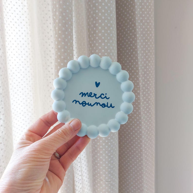 May include: A light blue, round coaster with a beaded edge. The coaster features the words "merci nounou" in dark blue script, with a small heart above the text. The coaster is held in a hand.