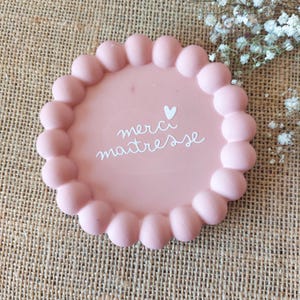 May include: A pink, round decorative tray with a scalloped edge. The tray features the words "merci maitresse" in white script, along with a small heart. The tray is on a textured, tan surface.
