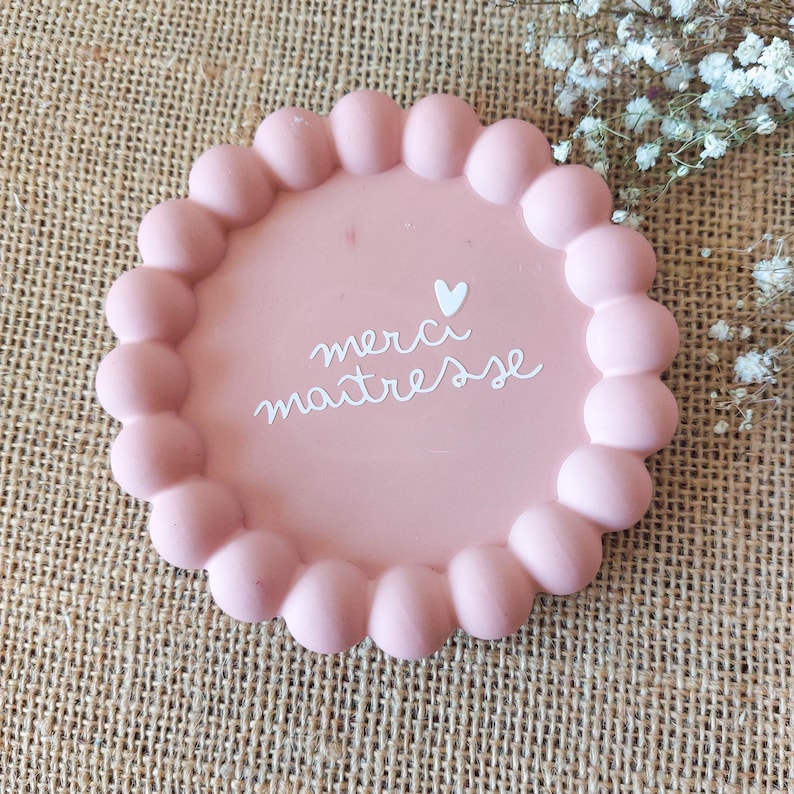 May include: A pink, round decorative tray with a scalloped edge. The tray features the words "merci maitresse" in white script, along with a small heart. The tray is on a textured, tan surface.