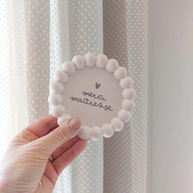 May include: A white, round coaster with a beaded edge is held in a hand. The coaster has the words "merci ma&icirc;tresse" and a heart symbol in the centre. The background features a polka dot curtain.