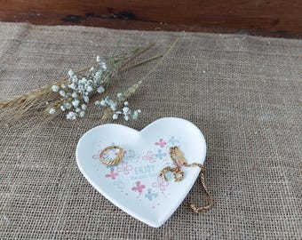 Może przedstawiać: Biała ceramiczna serduszko-kształtna miseczka z kwiatowym wzorem i napisem "Enjoy the little things". Miseczka leży na powierzchni z juty i zawiera złoty pierścionek i bransoletkę.