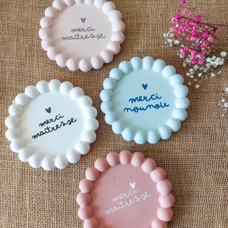 May include: Four pastel-coloured coasters with scalloped edges. The coasters are pink, white, and light blue. Each coaster features a handwritten French message, including "merci maitresse" and "merci nounou", along with a small heart symbol.