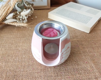 May include: A pink and white marbled wax warmer with a silver dish holding a pink wax melt. A lit tea light candle sits below. The warmer is on a burlap surface, with a book and dried flowers in the background.