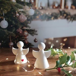 Może przedstawiać: Dwie białe ceramiczne figurki aniołów z czerwonymi sercami na piersiach, stojące na drewnianej powierzchni z zielonym gałązką ostrokrzewu i białymi światełkami choinkowymi.