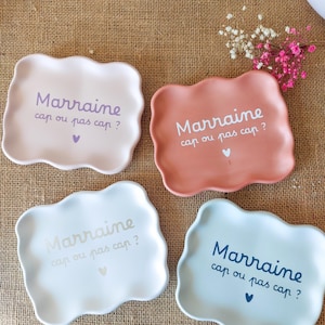 Può includere: Quattro piattini rettangolari in ceramica con bordi ondulati nei colori lilla chiaro, terracotta, bianco e azzurro. Ogni piattino presenta la scritta "Marraine cap ou pas cap ?" in un colore coordinato, con una piccola icona a forma di cuore.