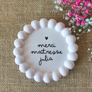 May include: A white, round decorative plate with a scalloped edge. The plate features the words "merci ma&icirc;tresse julia" in black script, with a small black heart above the text. The plate is on a burlap surface, with pink and white flowers in the background.