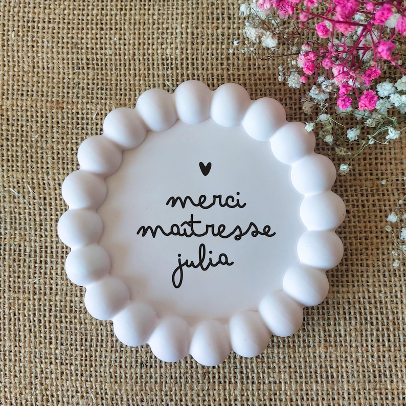 May include: A white, round decorative plate with a scalloped edge. The plate features the words "merci ma&icirc;tresse julia" in black script, with a small black heart above the text. The plate is on a burlap surface, with pink and white flowers in the background.