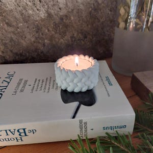 Könnte beinhalten: Weißer Kerzenhalter aus Keramik mit geflochtenem Design. Der Halter ist mit einer kleinen weißen Teelichtkerze gefüllt, die brennt.