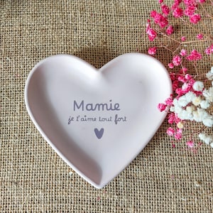 Peut inclure: Un vide-poche rose en forme de cœur avec l'inscription "Mamie je t'aime tout fort" en gris. Un petit cœur violet est en dessous du texte. Le plat est posé sur une surface en toile de jute, avec des fleurs roses et blanches.