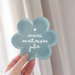 Może przedstawiać: Jasnoniebieski, dekoracyjny przedmiot w kształcie kwiatu z napisem "merci maitresse julia" w białym skrypcie. Małe białe serce znajduje się nad tekstem. Przedmiot jest trzymany w dłoni.