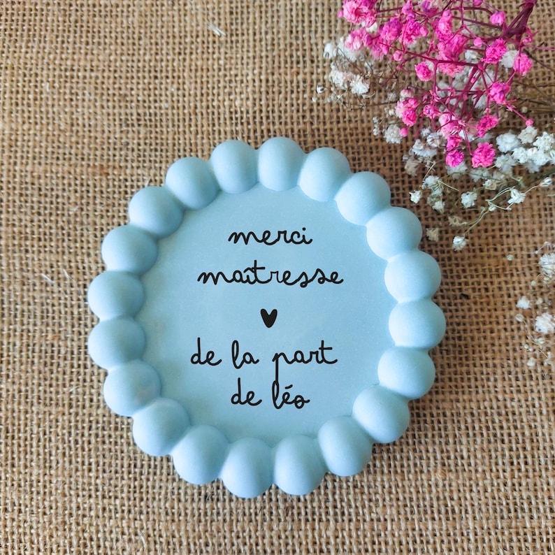 May include: A light blue, round decorative plaque with a scalloped edge. The plaque has the words "merci ma&icirc;tresse" and "de la part de L&eacute;o" written in black script. A small black heart is between the phrases. The background is a burlap texture.