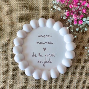May include: A white, round decorative plate with a scalloped edge. The plate has the words "merci nounou de la part de jade" in a brown script font. A small pink heart is between "nounou" and "de la part".