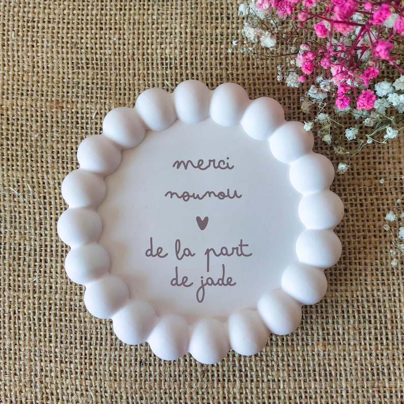 May include: A white, round decorative plate with a scalloped edge. The plate has the words "merci nounou de la part de jade" in a brown script font. A small pink heart is between "nounou" and "de la part".
