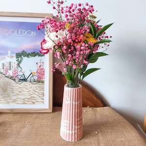 Può includere: Un vaso rosa con un design strutturato a linee verticali contiene un vivace bouquet di fiori rosa, gialli e bianchi con foglie verdi. Il vaso è appoggiato su una superficie marrone, con una stampa incorniciata di una bicicletta e un edificio sullo sfondo.