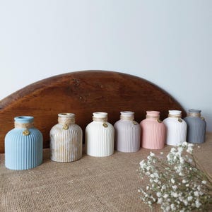 Op de afbeelding: Een set van acht kleine, decoratieve vazen in verschillende kleuren, waaronder lichtblauw, beige, wit, grijs en roze. Elke vaas heeft een geribbelde textuur en een klein gouden accent.