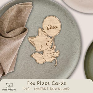 Op de afbeelding: Houten plaatskaart in de vorm van een vos die een ballon vasthoudt met de naam "Oliver". De vos is lichtbruin en heeft een cartoonachtig ontwerp. De plaatskaart ligt op een grijze plaat met een gevouwen beige servet.