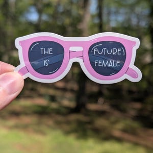 Può includere: Sticker di occhiali da sole rosa con il testo "The future is female" in lettere bianche.