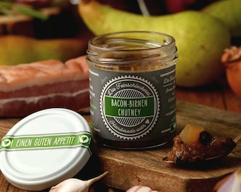 May include: A glass jar of bacon and pear chutney with a green label that reads "Die Feinschmeckerei Bacon-Birnen Chutney Handmade with". The jar is sitting on a wooden surface with a spoon of chutney, a clove of garlic, and pieces of bacon.