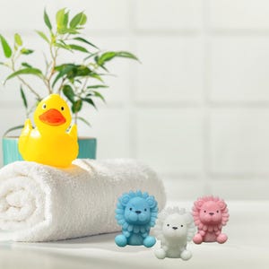 May include: A yellow rubber duck with an orange beak sits atop a rolled white towel. Three small lion-shaped soaps in blue, white, and pink are in front of the towel. A green plant in a blue pot is in the background.