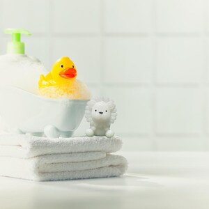 May include: A white bathtub filled with foamy bubbles, a yellow rubber duck with an orange beak, and a white lion figurine sit on a stack of white towels. A bottle of soap with a green pump is in the background. The scene is set against a white tiled wall.