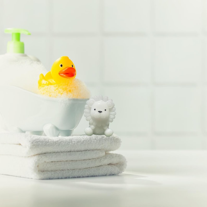 May include: A white bathtub filled with foamy bubbles, a yellow rubber duck with an orange beak, and a white lion figurine sit on a stack of white towels. A bottle of soap with a green pump is in the background. The scene is set against a white tiled wall.