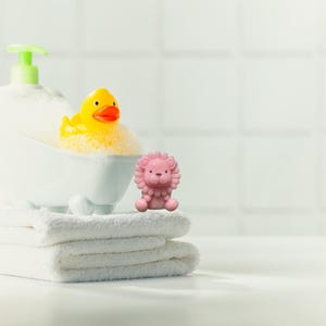 May include: A yellow rubber duck with an orange beak sits in a miniature white bathtub filled with bubbles. A pink lion figurine sits on a stack of white towels. A bottle of soap with a green pump is also visible.
