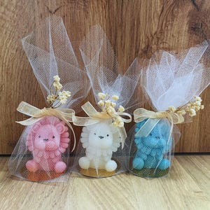 May include: Three lion-shaped soaps in pink, white, and blue, each wrapped in sheer white netting and tied with a gold ribbon and dried flowers. The soaps sit on a wooden surface.