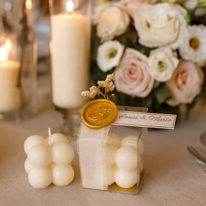 Puede incluir: Una decoración de mesa de boda con un ramo de rosas blancas y rosas, velas y una pequeña caja de regalo. La caja de regalo contiene velas de burbujas blancas y está sellada con un sello de cera dorado. Una pequeña tarjeta dice "Louis & Marie."