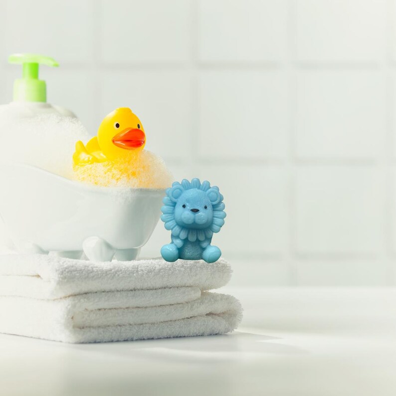 May include: A yellow rubber duck in a white bathtub filled with bubbles, next to a blue lion figurine. The bathtub sits on a stack of white towels, with a white soap dispenser in the background. The scene is set in a bathroom.