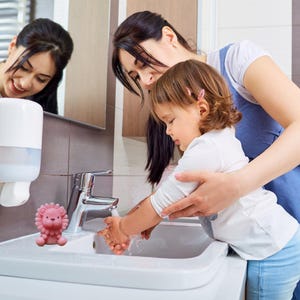 May include: A pink lion-shaped soap sits on the edge of a white sink. A child washes their hands with assistance from an adult. A mirror reflects the scene. The bathroom has a neutral color scheme.
